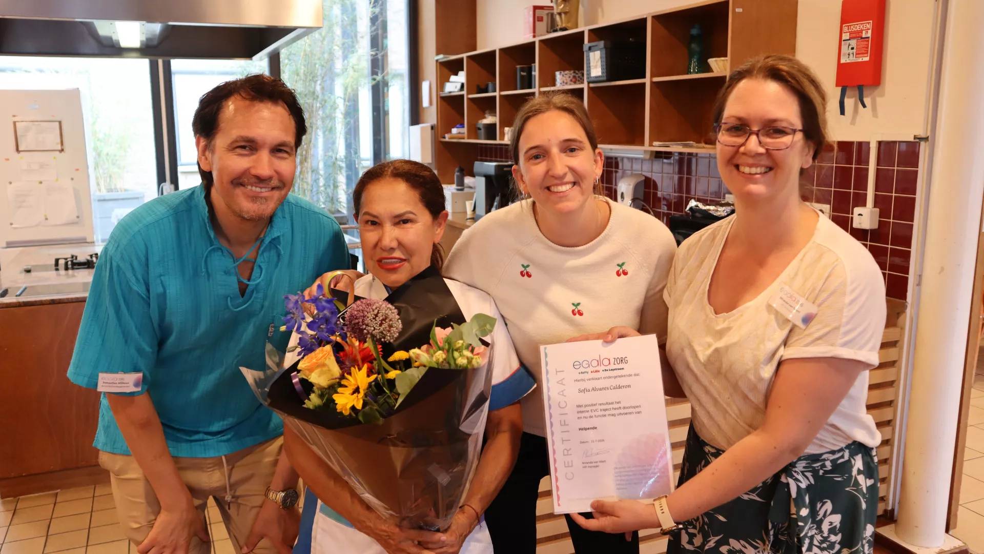 Een groep mensen poseert met bloemen en een certificaat in een keuken.