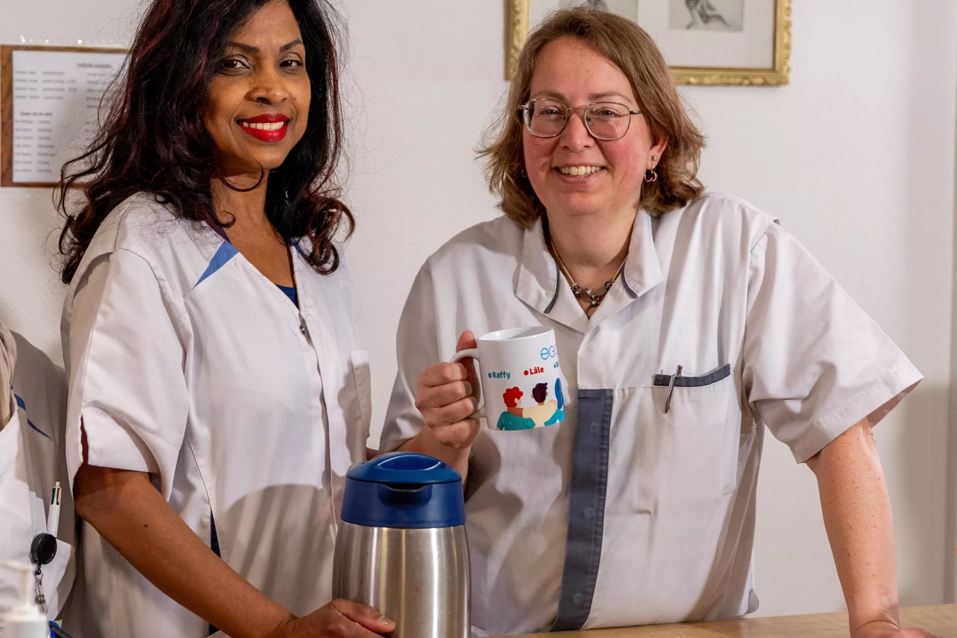 Twee medewerkers in uniform staan bij een balie met een thermoskan en mok.